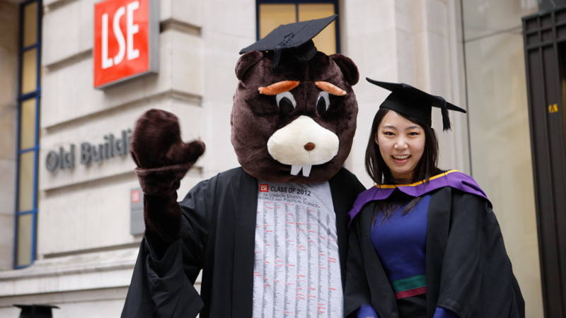 beaver waving with graduand in gown outside the Old building on Houghton Street