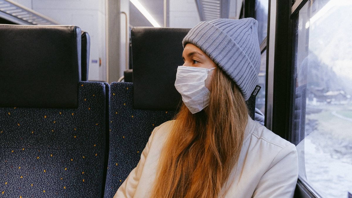 A girl sits on a train next to a window wearing a facemask and a grey hat