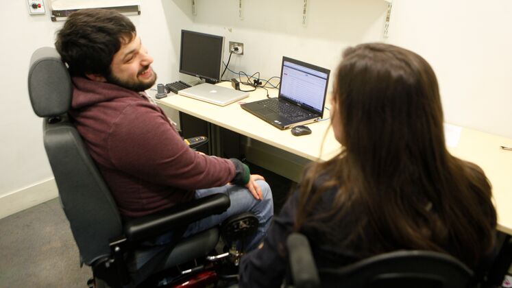 A man with a disability sitting at a lap top chatting to his academic mentor.