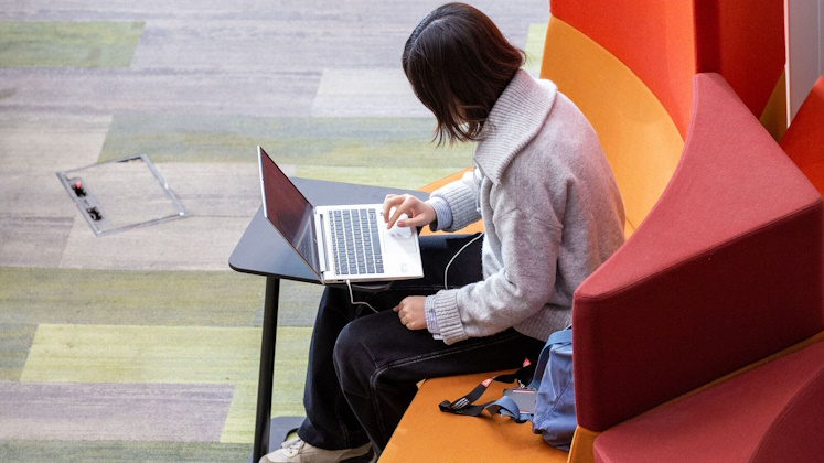 A student sits on a comfy chair inside a university building browsing on their laptop.