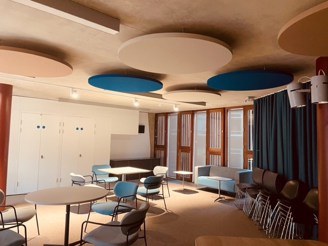 A closer look at the Community Space and the seating available. Small tables are shown, surrounded by blue and grey chairs. Next to these is a blue and grey modern sofa, and extra plastic chairs are stacked up next to this.