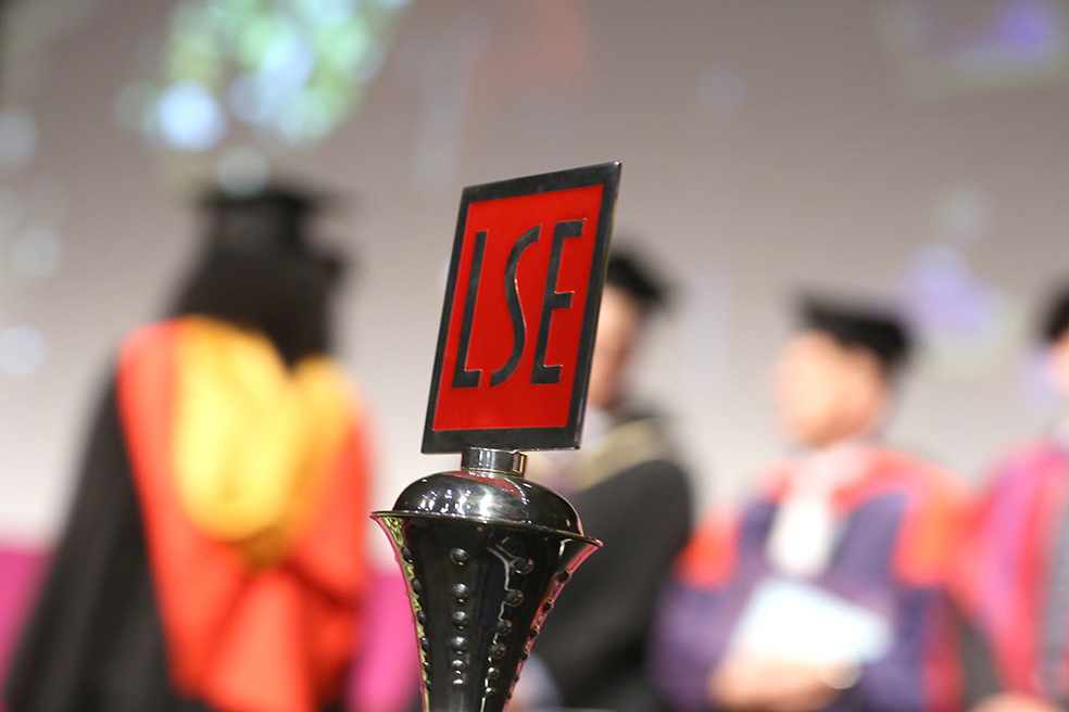 The LSE logo in metal on a bottle stopper, with students graduating in the background